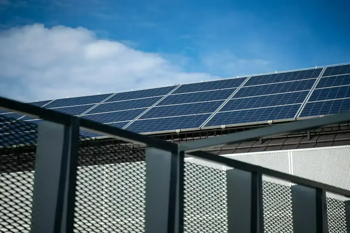Painéis solares em telhado sob céu azul com nuvens, com grade e parede perfurada em primeiro plano