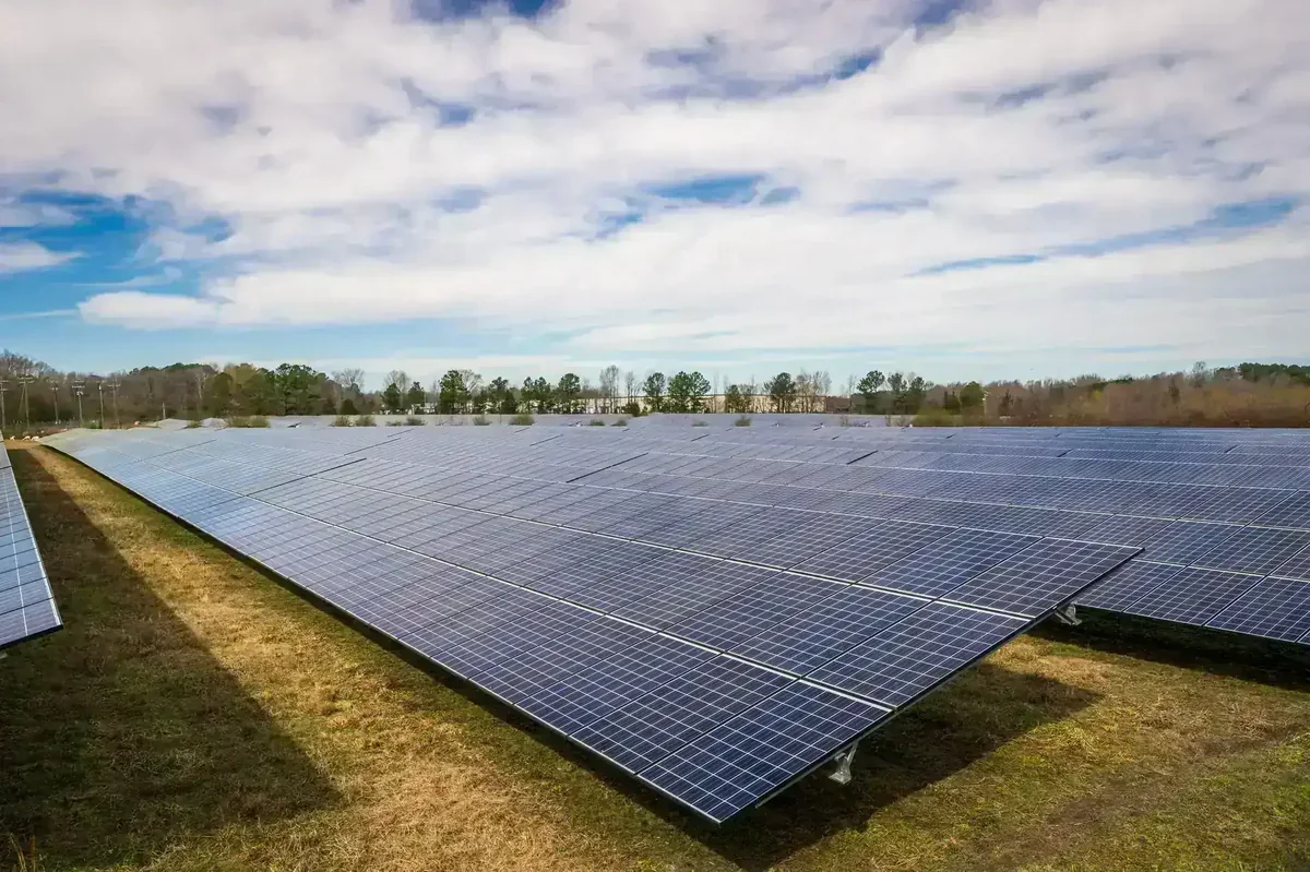 Extensa usina solar com painéis fotovoltaicos em campo aberto, céu azul com nuvens e árvores ao fundo
