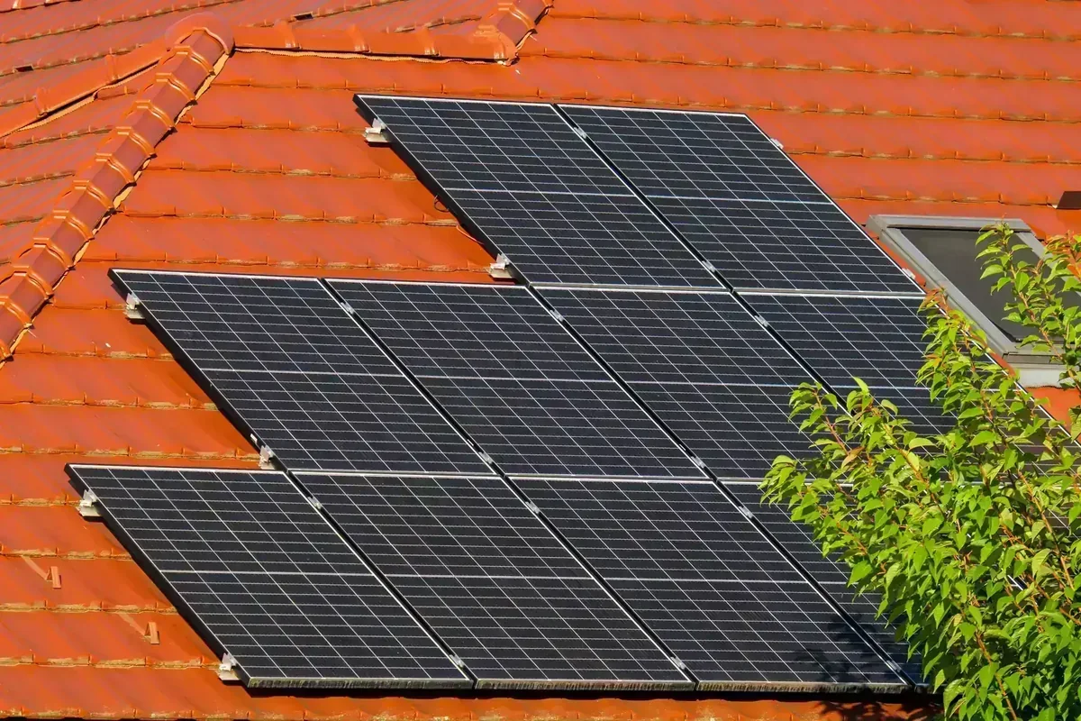 Painéis solares pretos instalados em telhado de telhas vermelhas, com claraboia e folhagem verde