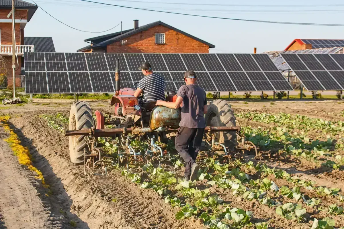 Homens cultivam lavoura com trator, com painéis solares de energia fotovoltaica e casas ao fundo