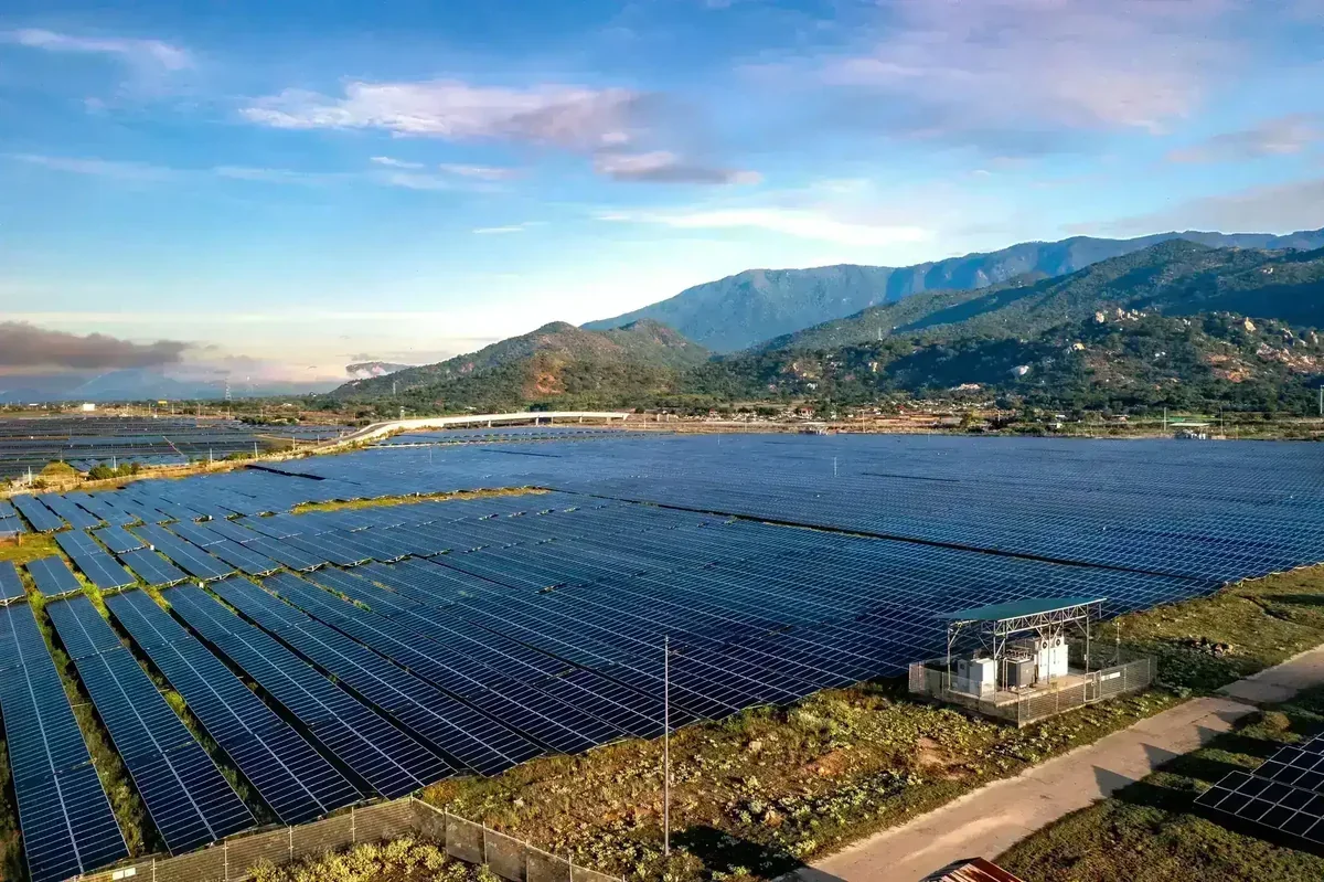 Vasta usina solar com painéis fotovoltaicos, montanhas e uma ponte ao fundo sob céu azul
