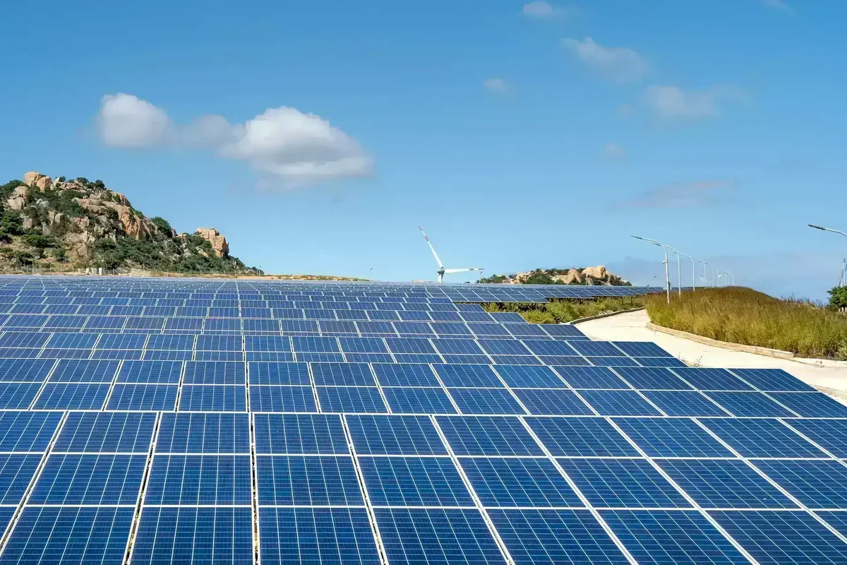 Campo de painéis solares azuis sob céu azul, com turbina eólica, morros rochosos e estrada com postes de luz
