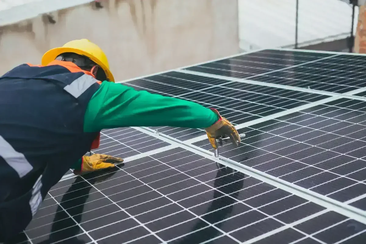 Técnico com capacete amarelo e luvas instalando painéis solares pretos em um telhado