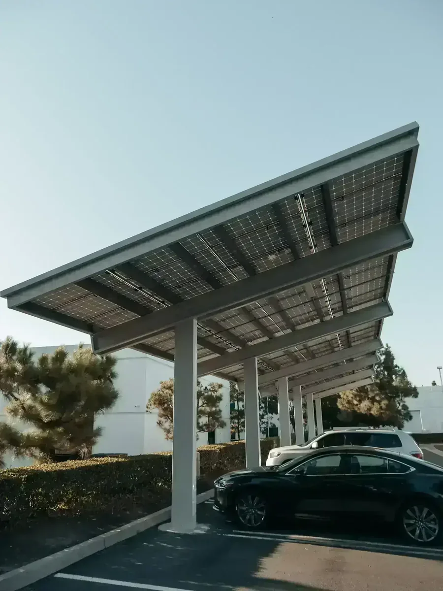 Carport solar com painéis fotovoltaicos cobrindo carros em estacionamento. Edifícios e árvores ao fundo